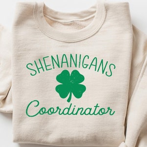 May include: A beige sweatshirt with the text "Shenanigans Coordinator" and a green four-leaf clover graphic.