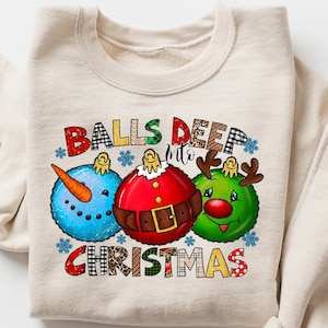 以下が含まれることがあります： 「BALLS DEEP INTO CHRISTMAS」と書かれたカラフルなグラフィックが特徴のベージュのスウェットシャツ。グラフィックは、雪だるま、サンタ、トナカイの3つのクリスマスオーナメントキャラクターを特徴としています。スウェットシャツは折りたたまれています。