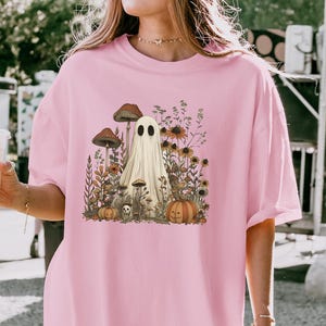 May include: A light pink t-shirt featuring a whimsical illustration of a ghost surrounded by mushrooms, pumpkins, and flowers. The design has a vintage, autumnal aesthetic. The shirt is a relaxed fit.