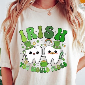 Może przedstawiać: Biały T-shirt z zielono-białym nadrukiem przedstawiającym dwie kresk&oacute;wkowe zęby w zielonych kapeluszach trzymające szczoteczkę do zęb&oacute;w i butelkę płynu do płukania ust. Pod zębami znajduje się napis "Irish You Would Floss".