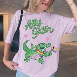 May include: A light purple t-shirt featuring a green cartoon alligator graphic with the text Ally Gator in green script. The alligator wears a rainbow pride bandana and holds a small rainbow flag. Small pink hearts surround the central illustration.