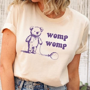Può includere: Una maglietta beige chiaro con un orsacchiotto di peluche viola che tiene un palloncino viola. Il testo "womp womp" è stampato in viola sotto l'orso.