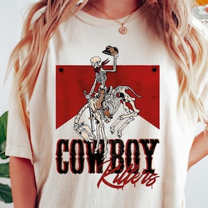 May include: A white t-shirt with a red and white graphic of a skeleton riding a horse. The graphic says "COWBOY Riders".
