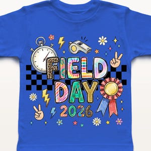 May include: A royal blue t-shirt featuring a vibrant graphic. The design includes the text "FIELD DAY 2026" in a tie-dye style, alongside a stopwatch, whistle, peace signs, and a ribbon. The shirt is likely for a school event.
