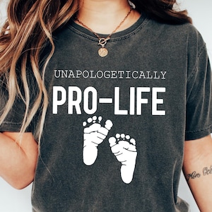 Puede incluir: Una camiseta gris oscuro con el texto "Unapologetically Pro-Life" y dos huellas de pies de bebé blancas. La camiseta la lleva puesta una persona con el pelo largo y castaño y un collar de oro.