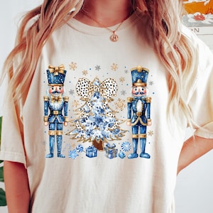 May include: A cream colored t-shirt with a blue and gold Christmas tree design featuring two nutcracker soldiers on either side of the tree. The tree has a white and black polka dot bow and is decorated with gold glitter and blue snowflakes.
