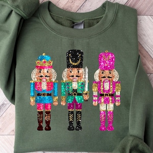 May include: A green sweatshirt featuring a festive design of three nutcracker soldiers. The nutcrackers are decorated with glitter and have gold accents.