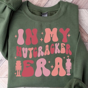 Puede incluir: Una sudadera verde con el texto "In My Nutcracker Era" en letras rosas y rojas con una silueta de cascanueces a cada lado.