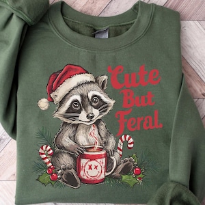 Pu&ograve; includere: Felpa verde natalizia con un adorabile procione che indossa un cappello da Babbo Natale e tiene una tazza di cioccolata calda. Il testo "Cute But Feral" &egrave; stampato sulla felpa.