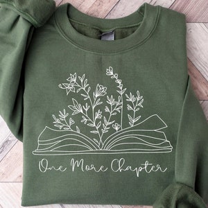 Peut inclure: Un sweat-shirt vert avec un motif blanc d'un livre ouvert avec des fleurs qui en poussent. Le texte "One More Chapter" est imprimé sous le livre.