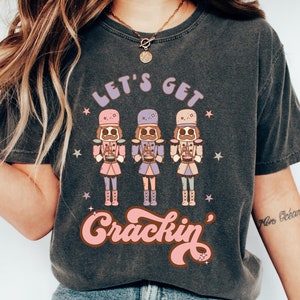 Puede incluir: Una camiseta gris oscuro con un gráfico de tres figuras de cascanueces tocando instrumentos. El texto "Let's Get Crackin'" está impreso en letras rosas y blancas.