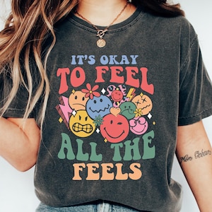 Op de afbeelding: Een donkergrijs T-shirt met de tekst "It's okay to feel all the feels" in kleurrijke letters. De tekst wordt omringd door een groep kleurrijke smiley-gezichten met verschillende uitdrukkingen.