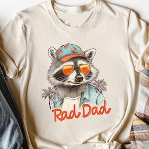 May include: Cream-colored t-shirt featuring a raccoon wearing sunglasses and a hat, with the text "Rad Dad" in red. The shirt is styled with a denim jacket, a plaid scarf, and a hat.