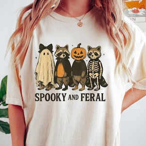May include: A cream-coloured t-shirt with a Halloween graphic. The design includes a ghost, two raccoons, a pumpkin-headed figure, and a skeleton raccoon. The text "SPOOKY AND FERAL" is printed beneath the characters.