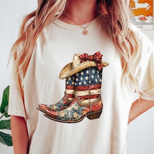 以下が含まれることがあります： クリーム色のTシャツ。カウボーイブーツと帽子のグラフィックが描かれており、アメリカ国旗のデザインで飾られています。ブーツは赤、白、青で、茶色のヒールと赤いリボンが付いています。シャツは人が着ています。