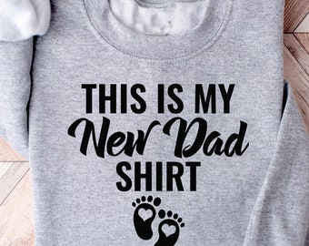 May include: Gray sweatshirt with the text "This is my New Dad Shirt" and a pair of black footprints with hearts on them.