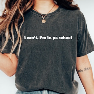 以下が含まれることがあります： 「i can't, i'm in pa school」の白い文字が入った濃いグレーのTシャツ。青いジーンズと白いスニーカーと合わせています。全体的にカジュアルでリラックスしたスタイルです。