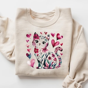 May include: A cream-colored sweatshirt featuring a watercolor illustration of a cat adorned with flowers and surrounded by pink and blue hearts. The cat's design includes floral patterns and heart accents, creating a whimsical and romantic theme.