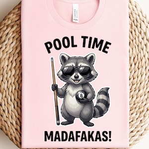 May include: A light pink t-shirt featuring a cartoon raccoon wearing sunglasses and holding a pool cue and an 8-ball. The text "POOL TIME MADAF*KAS!" is printed above and below the raccoon illustration.