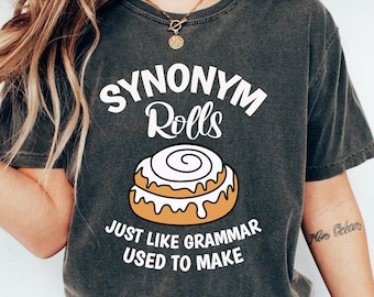 Può includere: Una maglietta grigia scura con il testo "Synonym Rolls" e un cartone animato di un cornetto alla cannella. Il testo sotto il cornetto dice "Proprio come la grammatica era solita fare."