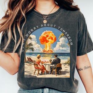 以下が含まれることがあります： レトロなイラストがプリントされた濃いグレーのTシャツ。ビーチサイドのテーブルでくつろぐカップルと、背景には核爆発が描かれています。「DON'T WORRY ABOUT TOMORROW」の文字がアーチ状に配置されています。柔らかい素材で作られています。