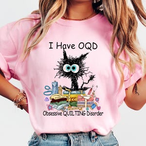 May include: Pink t-shirt with the text "I Have OQD" and "Obsessive QUILTING Disorder". The graphic features a black cat with large eyes sitting on a pile of quilting supplies, including fabric, scissors, and spools of thread.