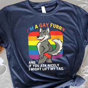 May include: Navy blue t-shirt with the text "I'M A GAY FURRY" and a cartoon wolf character. The wolf is standing in front of a rainbow-colored background. The text "AND IF YOU ASK NICELY I MIGHT LIFT MY TAIL" is below the wolf.