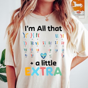 Op de afbeelding: Crèmekleurig T-shirt met de tekst "I'm All that + a little EXTRA" in zwarte en kleurrijke letters. Het shirt heeft een afbeelding van chromosomen en een hartsymbool, wat een uniek ontwerp creëert.