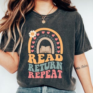 May include: A dark gray t-shirt with a rainbow, a book, and the text "READ RETURN REPEAT" in retro-style lettering.