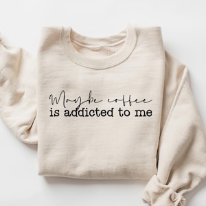 May include: Cream-colored sweatshirt with the text "Maybe coffee is addicted to me" in black script. The sleeves are rolled up, and the garment is laid flat. A casual, comfortable apparel item.
