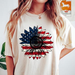 May include: Cream-colored t-shirt featuring a large sunflower design. The sunflower's petals are colored with the American flag, red and white stripes on the lower half and blue with white stars on the upper half. The shirt is a casual style.