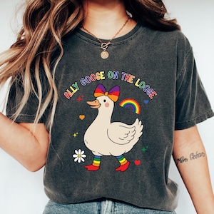 Puede incluir: Camiseta gris oscuro con un ganso de dibujos animados con un lazo arcoíris, calcetines arcoíris y un arcoíris. El texto "ALLY GOOSE ON THE LOOSE" está sobre el ganso. La camiseta es de estilo casual.