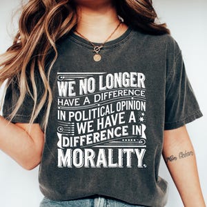 Op de afbeelding: Donkergrijs t-shirt met de witte tekst "WE NO LONGER HAVE A DIFFERENCE IN POLITICAL OPINION WE HAVE A DIFFERENCE IN MORALITY". De persoon draagt een blauwe jeans en witte schoenen. Het shirt heeft een casual stijl.