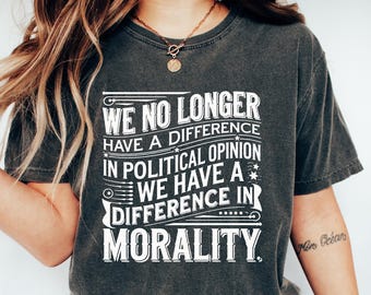 May include: Dark gray t-shirt with the text "WE NO LONGER HAVE A DIFFERENCE IN POLITICAL OPINION WE HAVE A DIFFERENCE IN MORALITY" in white. The person is wearing blue jeans and white shoes. The shirt is a casual style.