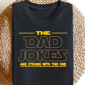 May include: Black t-shirt with yellow text that reads "THE DAD JOKES ARE STRONG WITH THIS ONE." The shirt is displayed on a woven, circular surface. The text is in a bold, sans-serif font.
