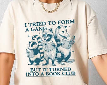 May include: A beige t-shirt with a graphic of three animals reading books. The text on the shirt reads "I tried to form a gang but it turned into a book club."