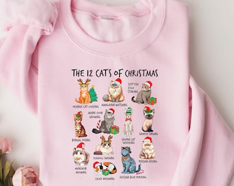 May include: Light pink sweatshirt with the text "The 12 Cats of Christmas" and illustrations of twelve different cats wearing Santa hats. Each cat is labeled with a humorous Christmas-themed name.