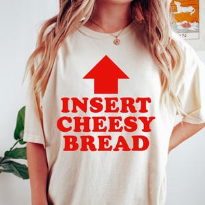 May include: Cream-colored t-shirt with the text "INSERT CHEESY BREAD" in red, topped with a red arrow pointing upwards. The shirt is worn with dark green shorts. A bracelet and necklace are also visible.