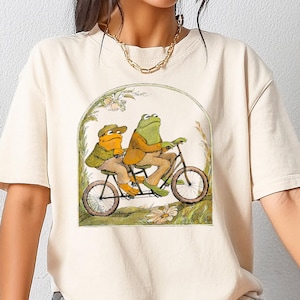 May include: A cream colored t-shirt with a graphic of two frogs riding a tandem bicycle. The frogs are wearing hats and the bicycle is surrounded by flowers.