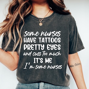 May include: Dark gray t-shirt with the text "Some nurses have tattoos pretty eyes and cuss too much. It's me, I'm some nurses" in white. The shirt is paired with blue jeans and white sneakers. A necklace and bracelet are also visible.