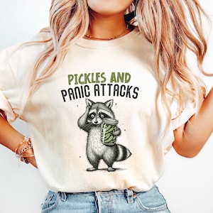 Könnte beinhalten: Cremefarbenes T-Shirt mit dem Text "Pickles and Panic Attacks" über einer Waschbär-Illustration. Der Waschbär hält ein Glas mit Essiggurken. Das Shirt wird mit hellblauen Jeans kombiniert.