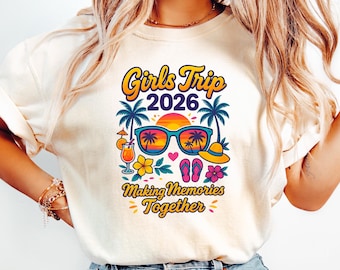 May include: Cream-colored t-shirt with a colorful graphic that reads "Girls Trip 2026 Making Memories Together." The design includes sunglasses, palm trees, a sunset, flowers, a cocktail, a hat, and flip-flops. The model is wearing light blue jeans.