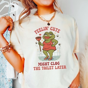 以下が含まれることがあります： 赤いドレスとハイヒールを履いた漫画のカエルのイラストが描かれたクリーム色のTシャツ。テキストは「Feelin' Cute Might Clog The Toilet Later」と赤とピンクで書かれています。シャツはブルージーンズと合わせて着用されています。