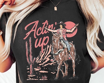 May include: Black t-shirt with a graphic of a cowboy riding a horse in a desert landscape. The text "Actin' up" is printed in pink on the shirt.