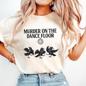 May include: Cream-colored t-shirt with the text "MURDER ON THE DANCE FLOOR" above a disco ball graphic and three black raven illustrations. The shirt is paired with light blue denim jeans.