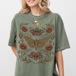 May include: Olive green t-shirt featuring a detailed illustration of three moths, surrounded by flowers and crescent moons. The design is in shades of brown, red, and blue. The shirt has short sleeves and a relaxed fit.