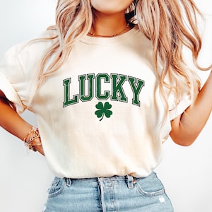 May include: Cream-coloured t-shirt featuring the word "LUCKY" in green block letters and a green shamrock. The shirt is worn with light blue denim jeans.