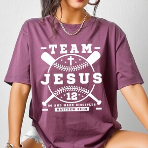 Puede incluir: Una camiseta granate con el texto "-TEAM- JESUS 12" en blanco, con un diseño de béisbol con bates cruzados y una cruz. La camiseta también incluye el texto "GO AND MAKE DISCIPLES MATTHEW 28:19".
