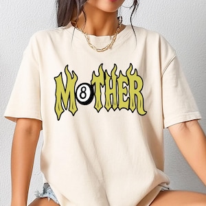 May include: Cream-colored t-shirt with the word "MOTHER" in a stylized, green and yellow font, featuring an eight-ball graphic. The shirt has short sleeves and a relaxed fit.