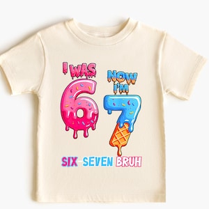 May include: Cream-colored youth t-shirt with a colorful graphic. The graphic reads "I WAS 6 NOW I'M 7 SIX SEVEN BRUH" with the numbers designed as dripping ice cream cones. The number 6 is pink, and the number 7 is blue and orange.